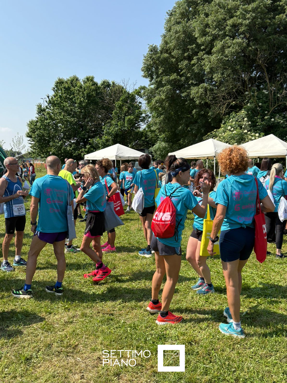 Partecipanti all’evento sportivo outdoor organizzato da Gelsia, riuniti in un’area verde sotto gazebo bianchi in una giornata di sole. I partecipanti indossano magliette personalizzate con il logo Gelsia, zainetti colorati e abbigliamento tecnico sportivo. Evento aziendale dedicato allo sport, al benessere e alla socializzazione.